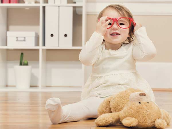 Little girl sitting on the floor with her teddy bear while playing with her glasses